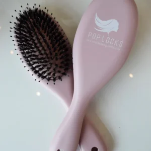Two pink Pop Locks Boar-Bristle Brushes with black bristles are crossed on a white surface. One brush features the POPLOCKS logo and "HAIR EXTENSIONS SPECIALIST," spotlighting Luxe Hair Studio, your Charleston hair salon.