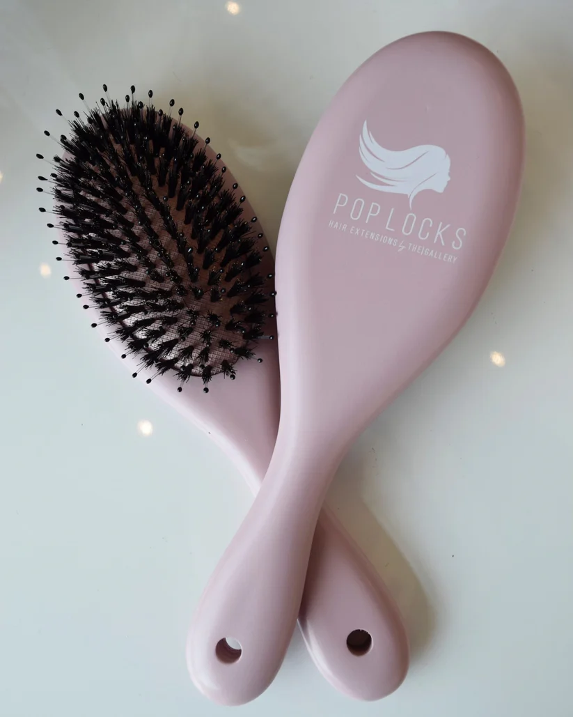 Two pink Pop Locks Boar-Bristle Brushes with black bristles are crossed on a white surface. One brush features the POPLOCKS logo and "HAIR EXTENSIONS SPECIALIST," spotlighting Luxe Hair Studio, your Charleston hair salon.