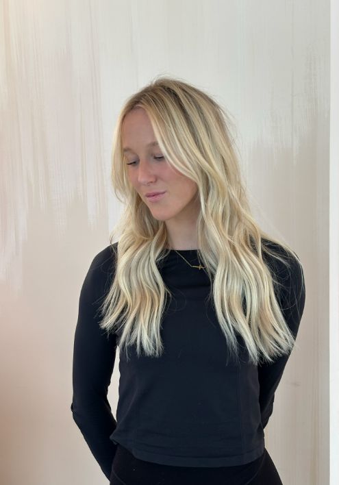A blonde woman with long hair stands elegantly in front of a textured wall, showcasing Luxe Hair Color.