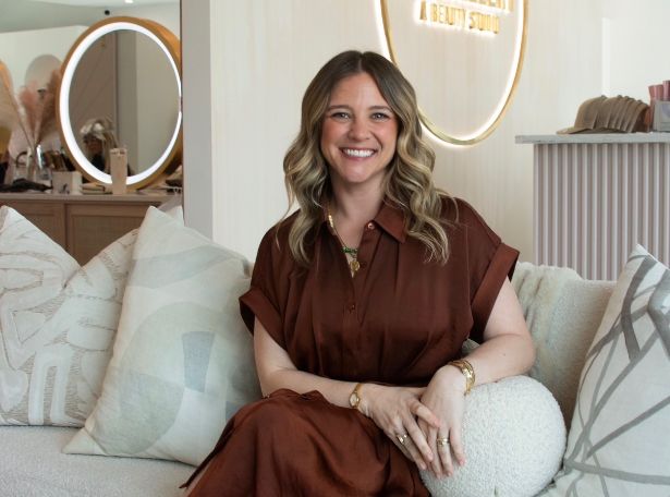 A woman with wavy, shoulder-length hair wearing a brown dress sits smiling on a couch with patterned cushions in a modern, well-lit beauty studio. A round mirror and decorative items are in the background.