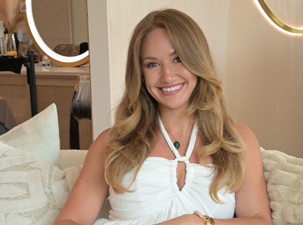 A woman with long, wavy blonde hair, wearing a white halter top, sits on a light-colored sofa and smiles at the camera. The setting appears modern and cozy, with soft lighting and neutral decor.