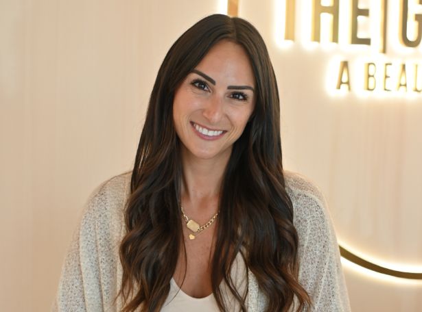 A woman with long, wavy brown hair and a warm smile stands indoors, wearing a light cardigan and gold necklace. There is a blurred sign with glowing letters in the background.