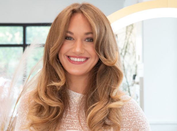 A woman with long, voluminous, wavy light brown hair and a bright smile wears a white textured top, standing in a well-lit modern room with soft natural and artificial lighting.