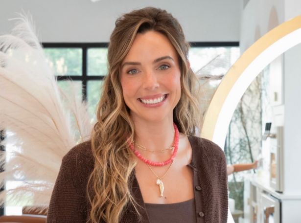 A woman with long wavy hair, wearing a brown sweater and layered necklaces, smiles indoors. The background includes plants, large windows, and modern decor.